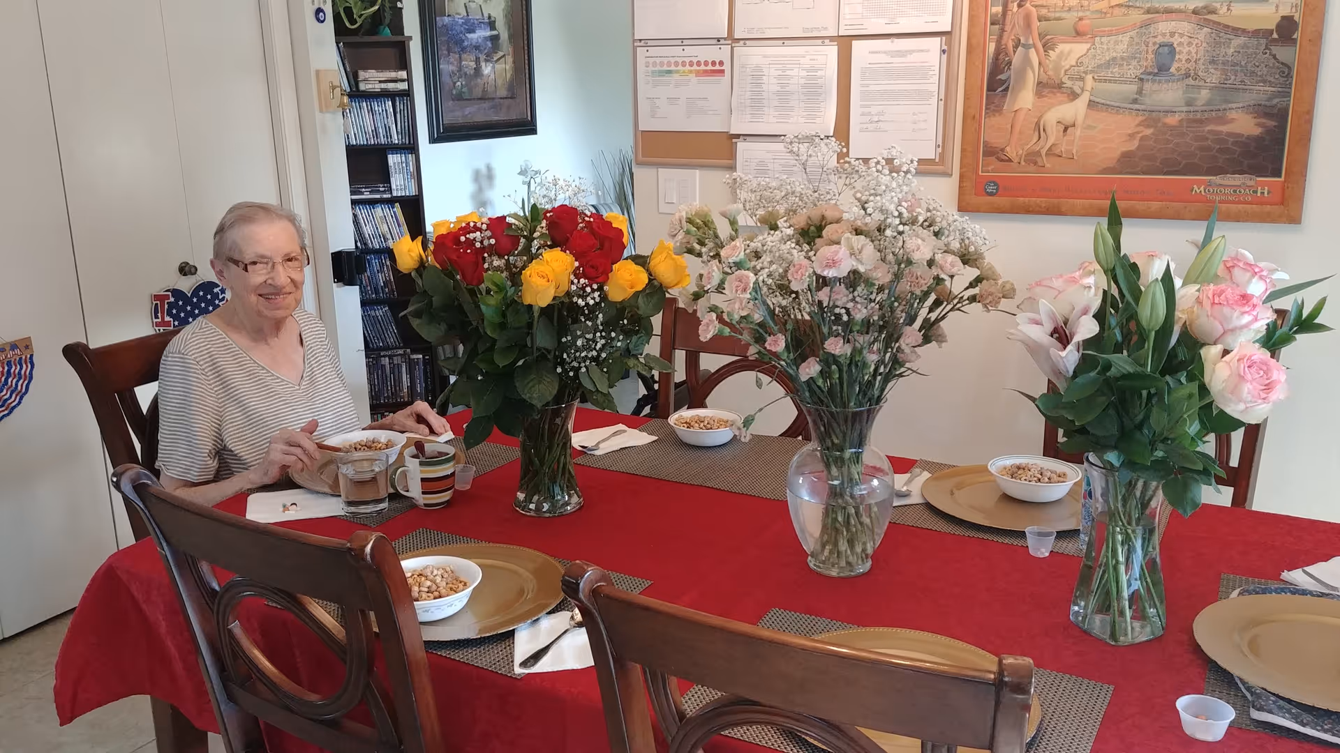 An elderly woman sits at a red-cloth dining table with flower arrangements.
