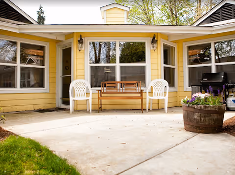 Front porch of a yellow building with white chairs, a wooden bench, and a planter.