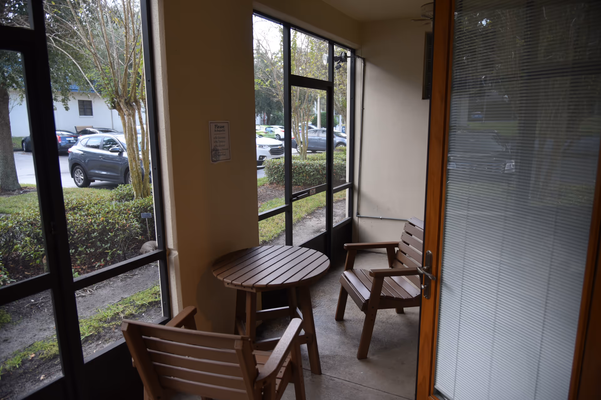 Wooden patio chairs and a round table sit inside a glass-enclosed balcony overlooking greenery.