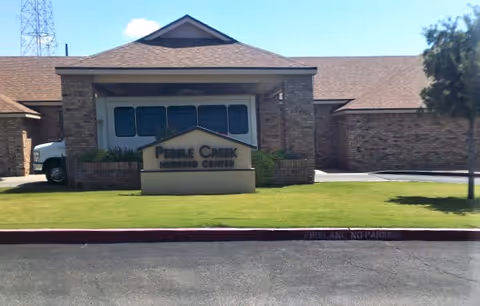 Front exterior view of Pebble Creek Nursing Center building with a sign displaying the facility name on a grassy area in front. The building has a brick facade and a covered entrance with a vehicle partially visible behind it.