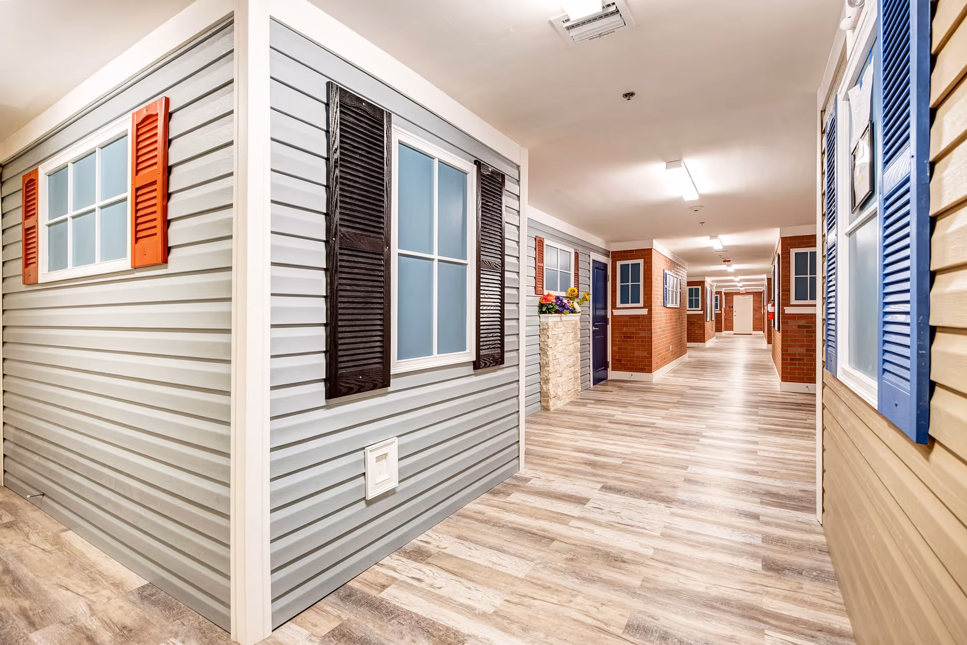 Interior corridor resembling a street with faux siding, shutters, and potted flowers.