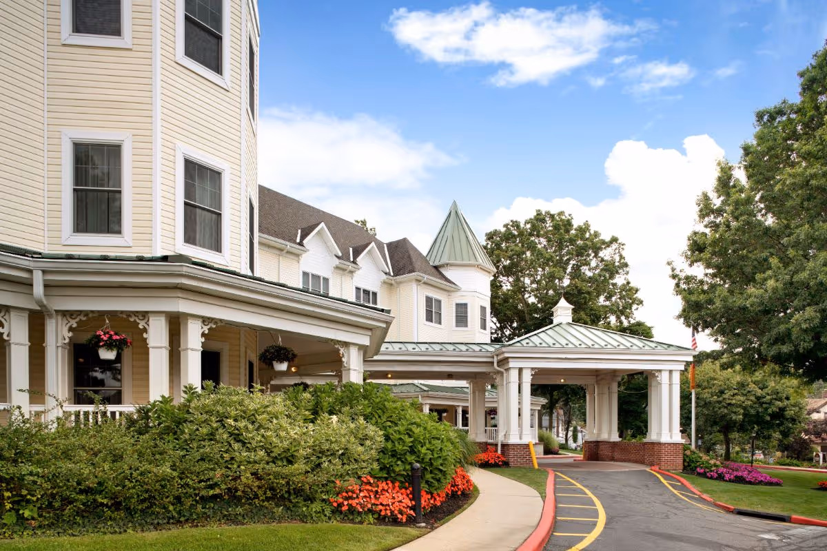 Exterior view of a senior living facility with a covered entrance, beige siding, white trim, and a green metal roof. The building is surrounded by well-maintained landscaping including bushes, flowers, and trees under a partly cloudy blue sky.