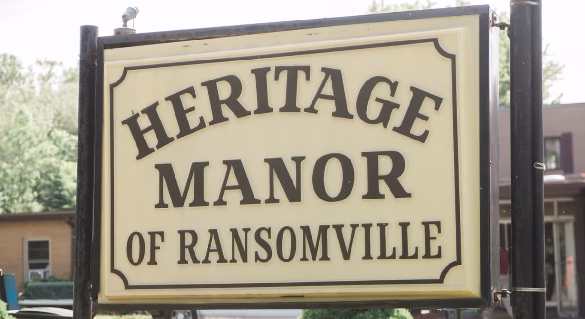 A sign reading Heritage Manor of Ransomville mounted outdoors beside a building.