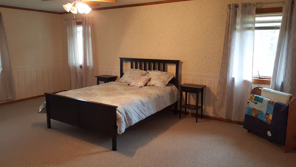 Bright, spacious bedroom with a dark wood bed, two nightstands, carpeted floor, and windows with sheer curtains.