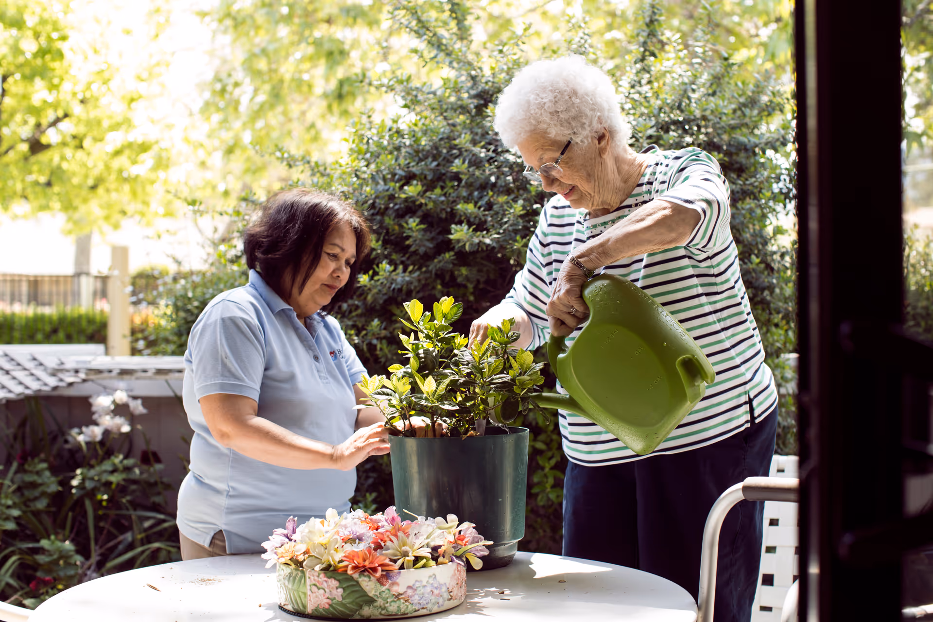 Two elderly women garden on an outdoor patio, watering plants beside a table with flowers.