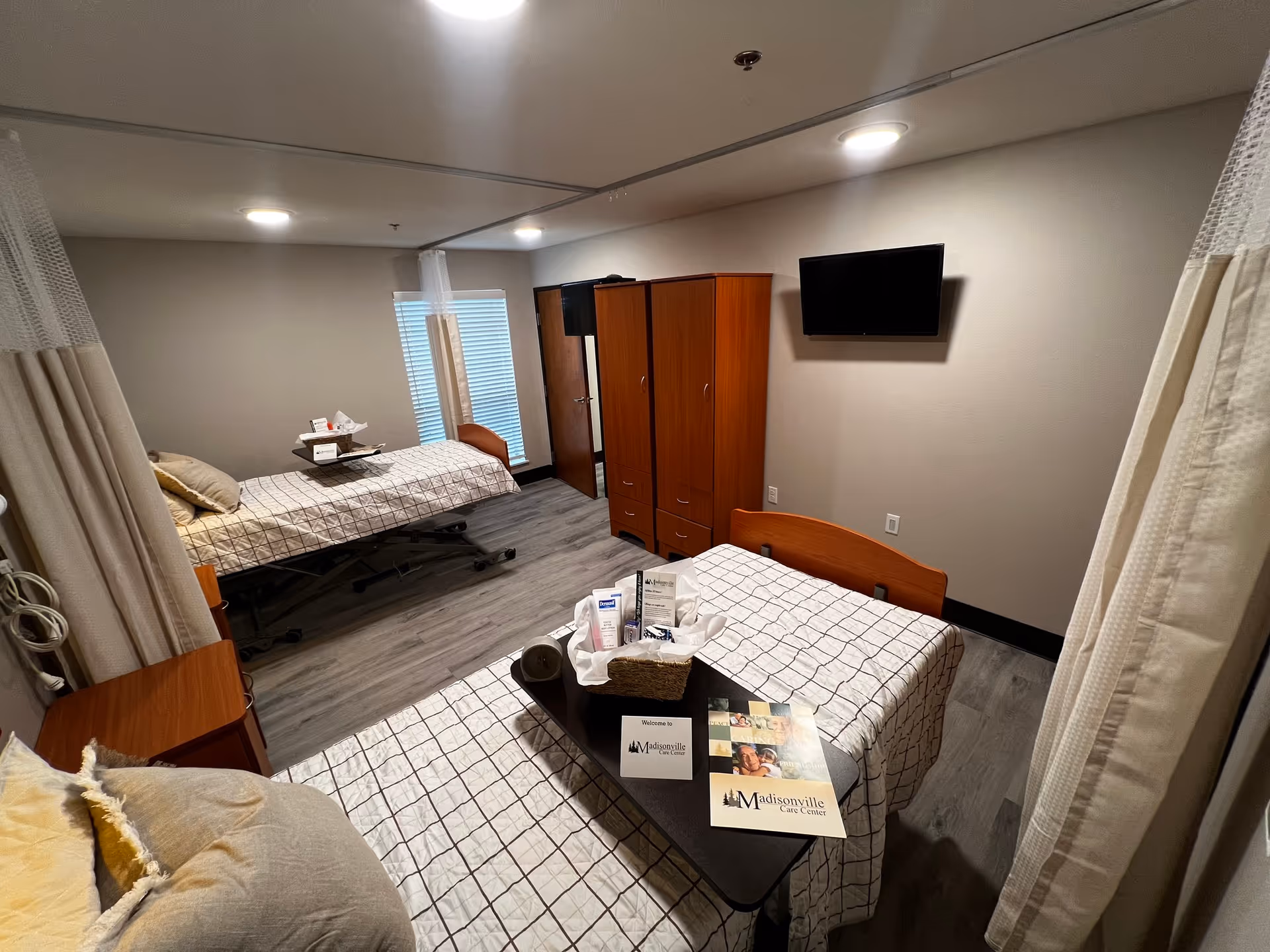 A shared patient room in a care facility with two hospital beds separated by privacy curtains. Each bed has a checkered bedspread and pillows. There is a small table on one bed with a basket containing toiletries and Madisonville Care Center brochures. The room has wooden wardrobes, a wall-mounted TV, and a window with blinds.