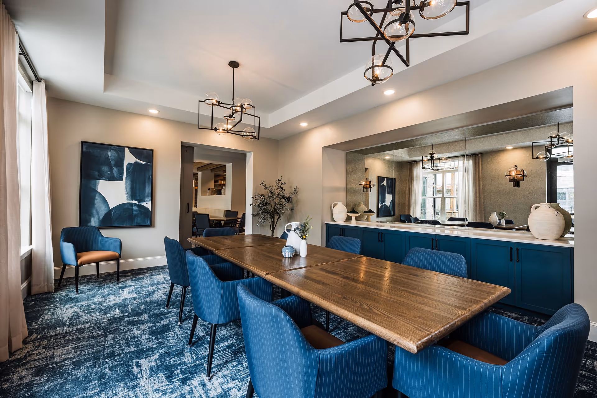 A modern dining room with a long wooden table surrounded by blue upholstered chairs. The room features a large mirror on one wall above blue cabinets, contemporary light fixtures hanging from the ceiling, a blue patterned carpet, and abstract artwork on the wall. Natural light enters through a window with sheer curtains.