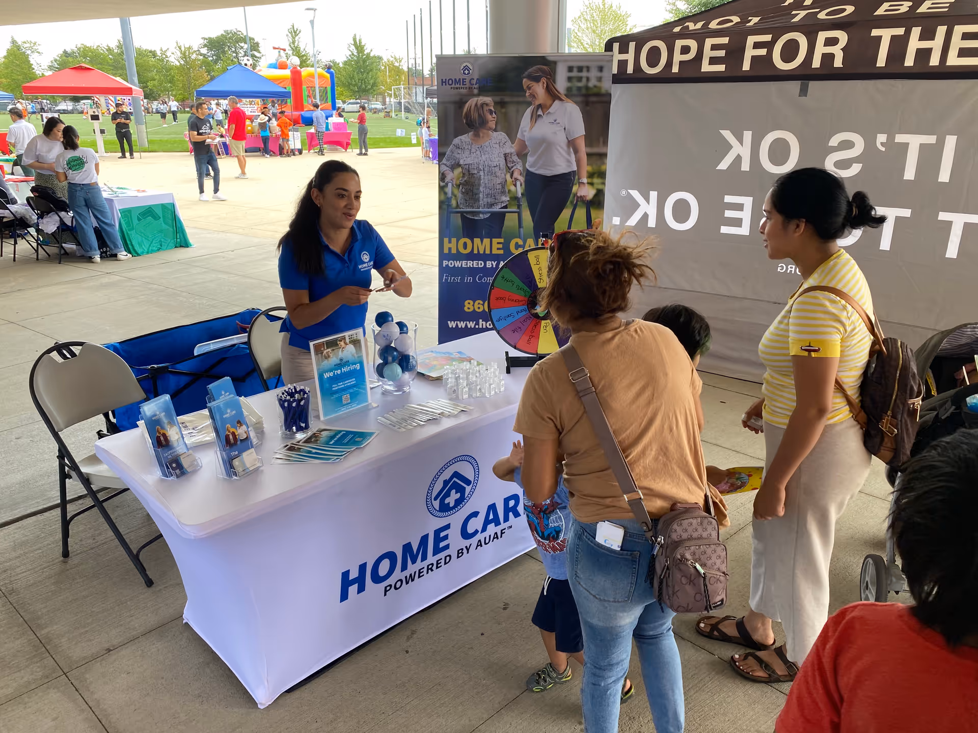 Blue-shirt caregiver speaks with attendees at an AUAF Home Care booth outdoors.