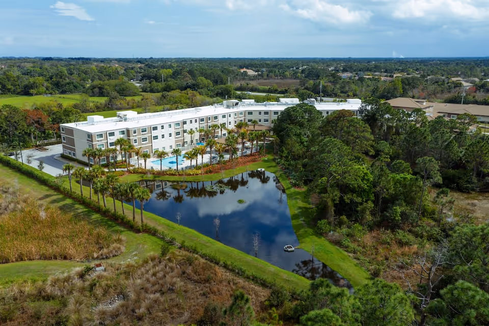 Aerial view of Discovery Village At Stuart, a large senior living facility surrounded by trees and greenery. The building is three stories tall with a white roof and beige exterior. There is a swimming pool visible near the center of the building, and a pond with reflections of the sky and trees in the foreground. The surrounding area includes grassy fields and wooded areas under a partly cloudy sky.