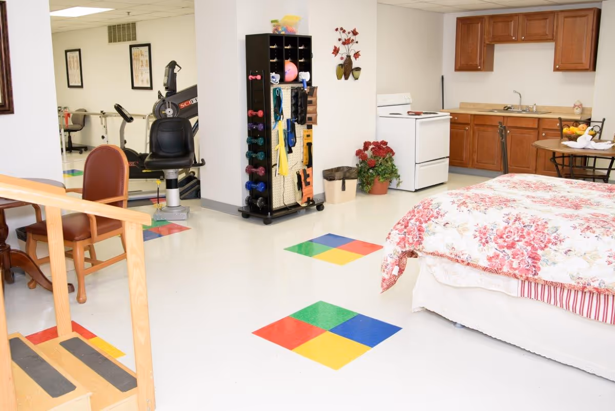 A bright room featuring a bed with a floral bedspread, a small kitchen area with wooden cabinets, a stove, and a round table with chairs. There is exercise equipment including a stationary bike and a rack with various weights and exercise accessories. The floor has colorful square patterns, and there are some potted plants and wall decorations.
