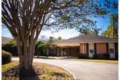 Brick senior living facility front with curved driveway, large tree, and manicured landscaping under blue sky.
