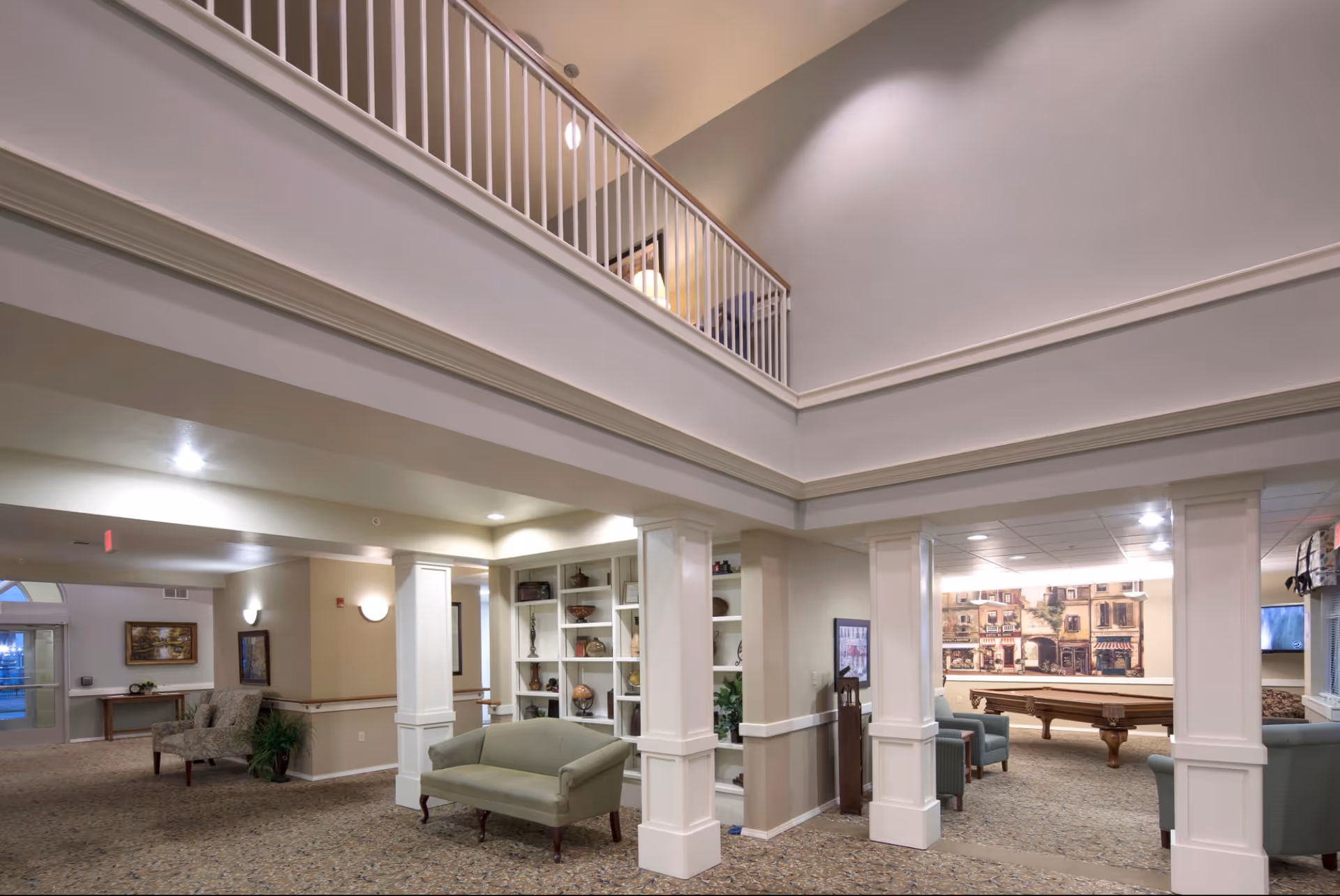Interior view of a senior living facility common area with a high ceiling and a second-floor balcony railing. The space includes a green upholstered sofa, built-in white shelves with decorative items, a pool table, and several armchairs. The walls are painted in neutral tones, and the carpet has a patterned design. There is a large mural of a street scene on one wall and framed artwork on other walls.