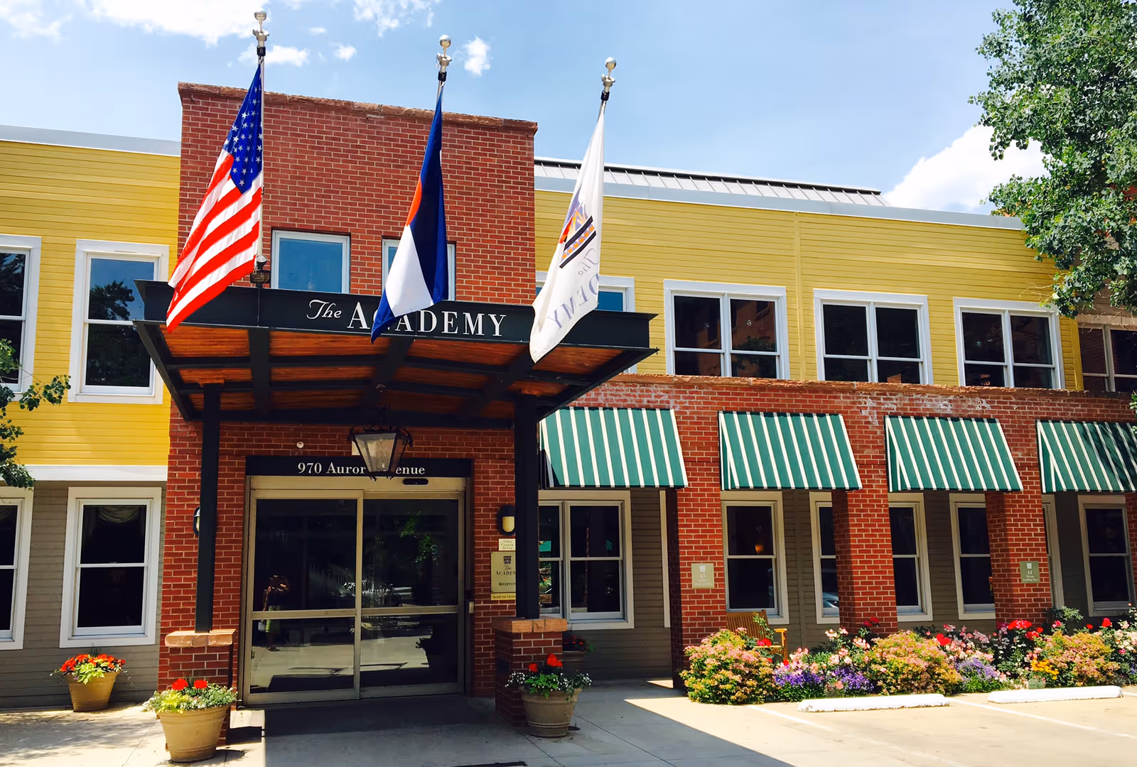 Front entrance of The Academy Boulder Communities building with flags, striped awnings, and flower planters.
