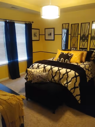 A decorated bedroom with a bed dressed in black-and-white patterned bedding, yellow accent pillows, black curtains, and a hanging ceiling light.
