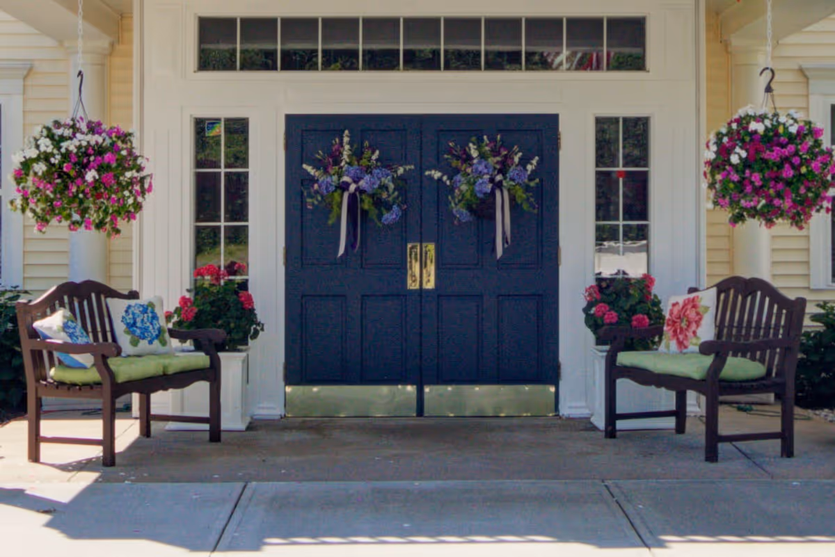 Two wooden chairs with green cushions flank a dark double front door adorned with floral wreaths
