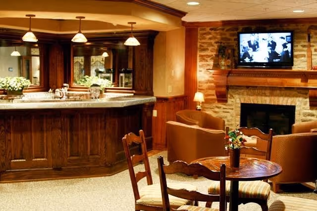 Cozy interior of a senior living facility featuring a wooden bar counter with pendant lights, a seating area with armchairs around a stone fireplace, and a wall-mounted TV above the fireplace. A small round wooden table with chairs and a flower vase is in the foreground.