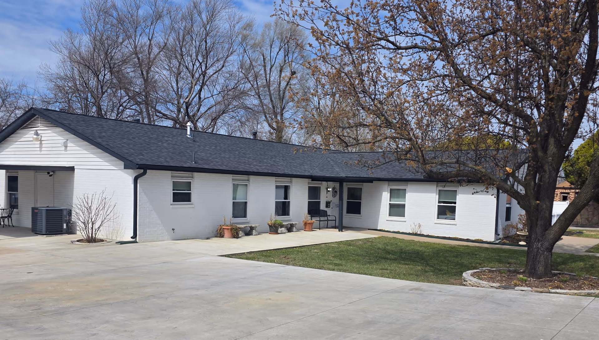 Single-story white facility with front entrance, lawn, and a large tree in the foreground.