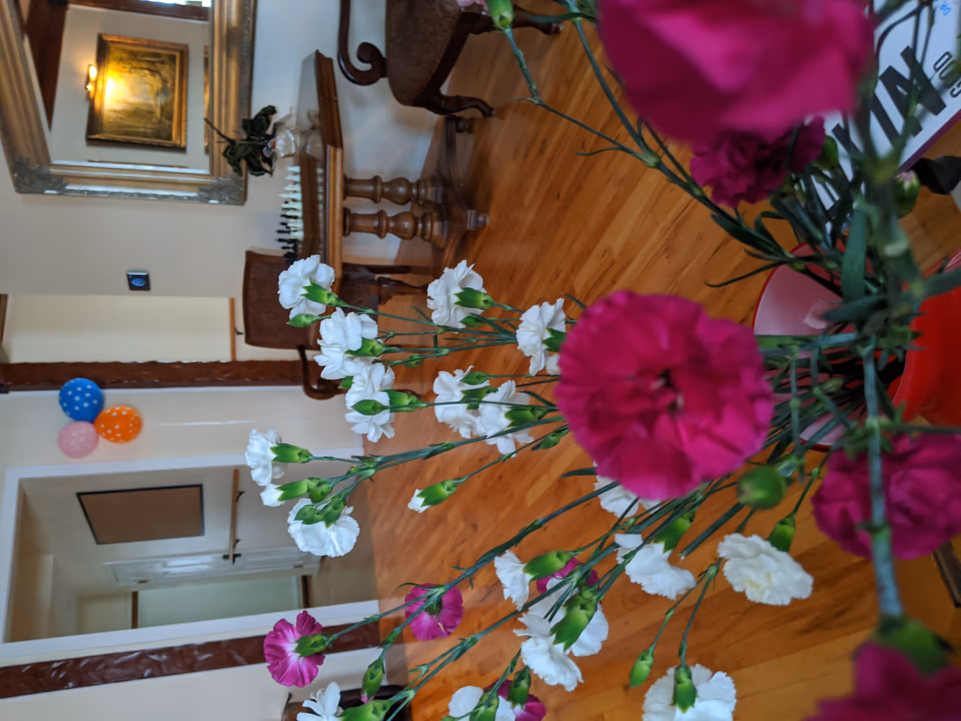 Bouquet of pink and white flowers in the foreground of a senior living facility common room with hardwood floors, a bench, mirror, and balloons on the wall.