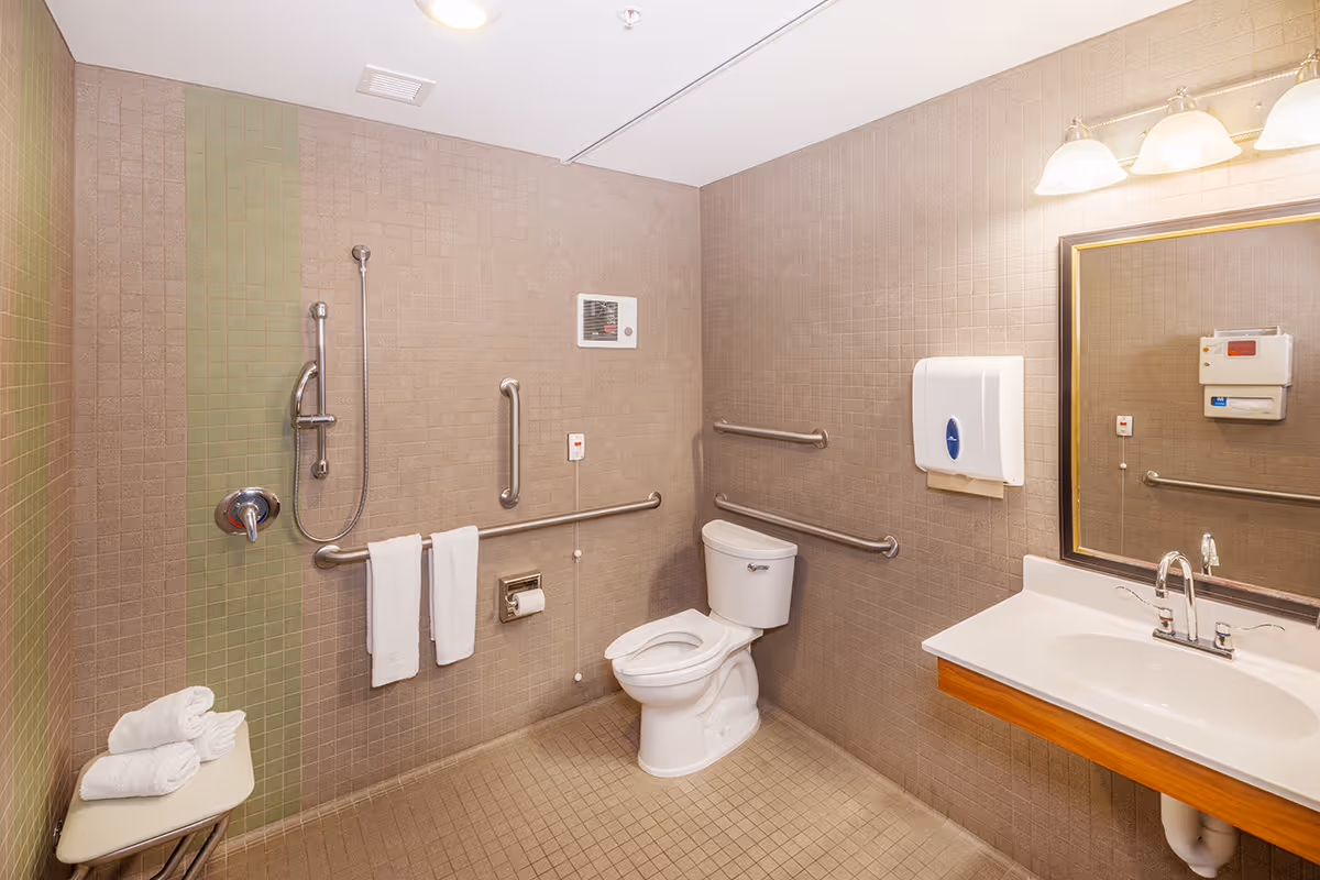 A clean, accessible bathroom with beige tiled walls and floor. It features a white toilet with grab bars on the walls around it, a shower area with a handheld showerhead and grab bars, a white sink with a large mirror above it, a wall-mounted soap dispenser, and a small bench with folded white towels.