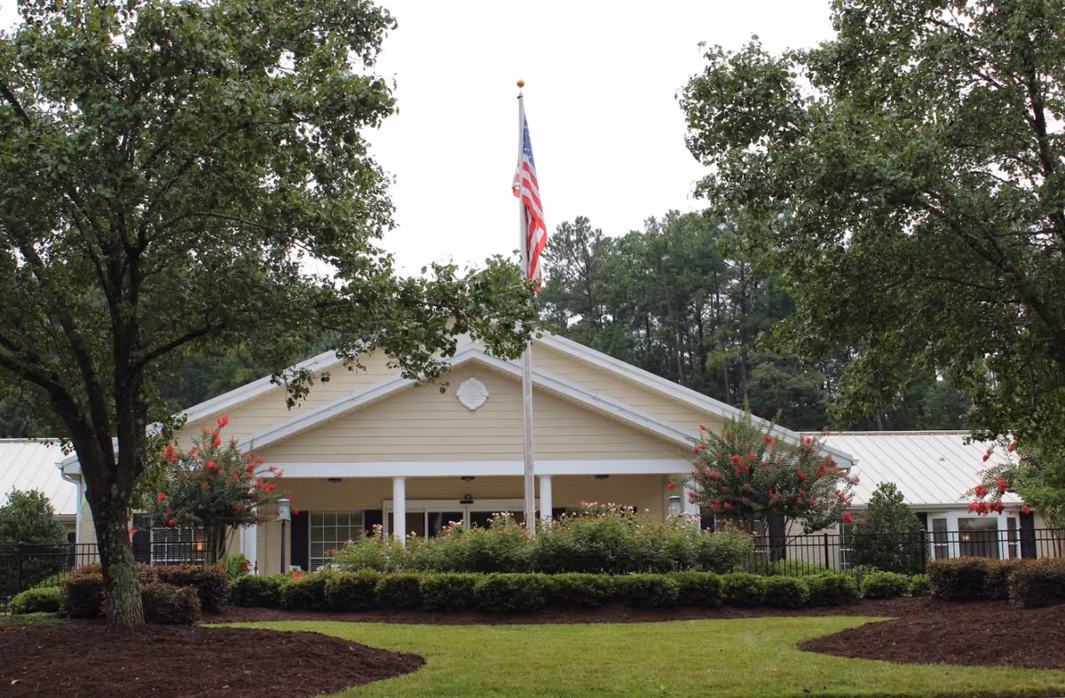 Front exterior view of a single-story building with a beige facade, white trim, and a metal roof. The building is partially obscured by trees and bushes, with an American flag on a flagpole in front. The surrounding area is landscaped with green grass, shrubs, and flowering plants.