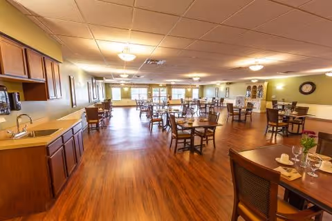 Spacious dining room with wooden flooring, multiple tables and chairs arranged neatly, soft yellow walls, ceiling lights, and a small kitchenette area with cabinets and a sink on the left side.