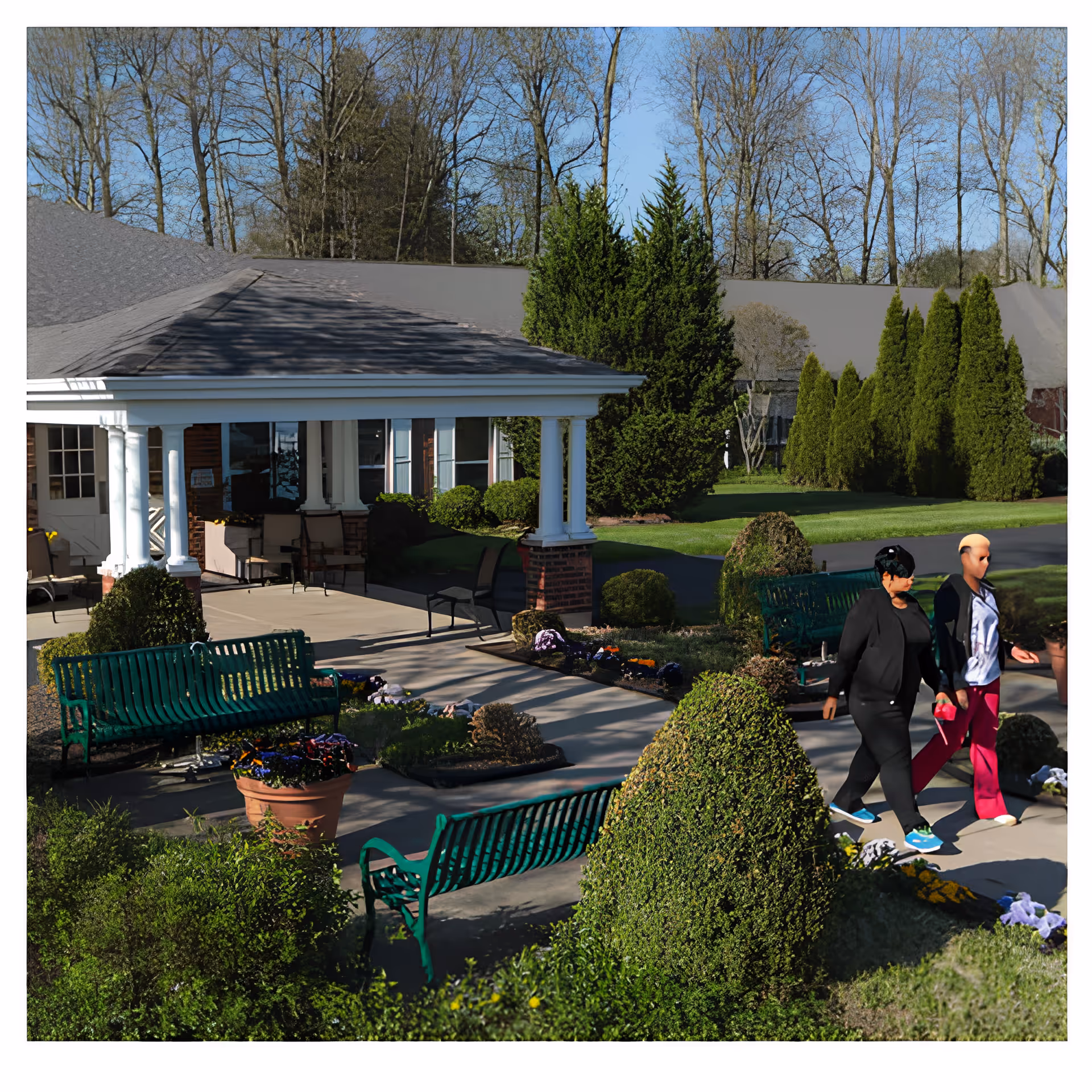 Front entrance and landscaped courtyard of a care facility with benches, potted flowers, and two people walking along the pathway.