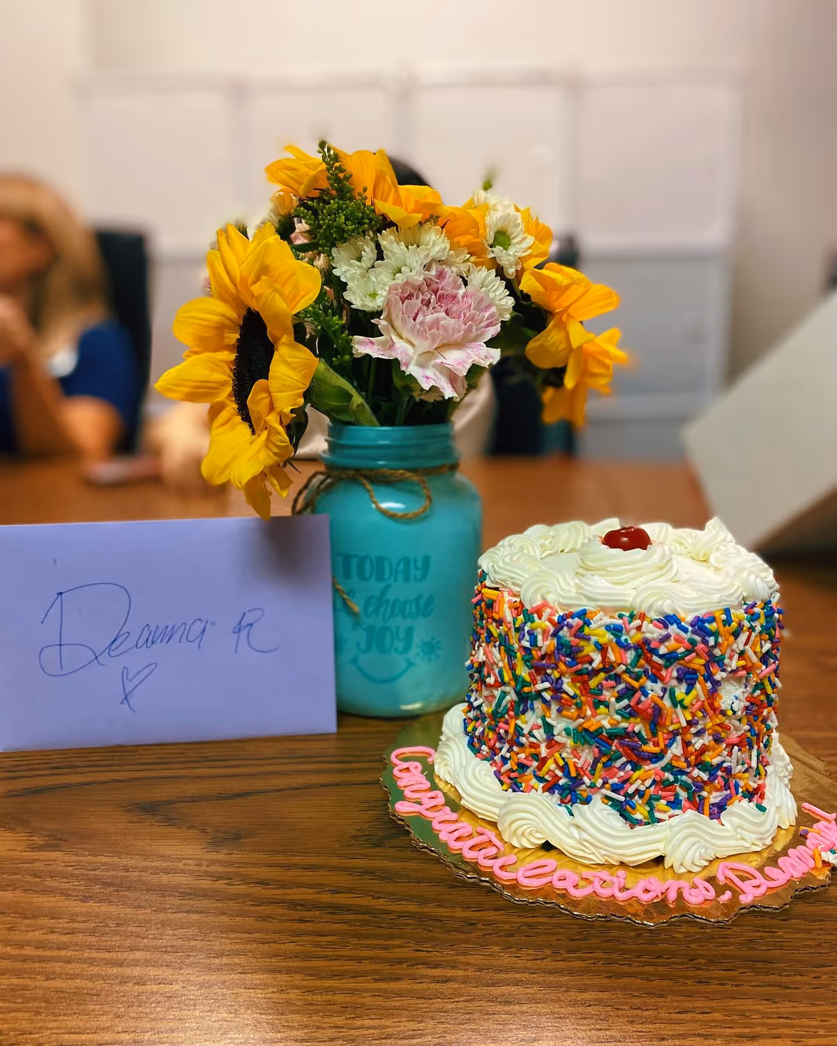 A cake with colorful sprinkles sits on a wooden table beside a bouquet of sunflowers.