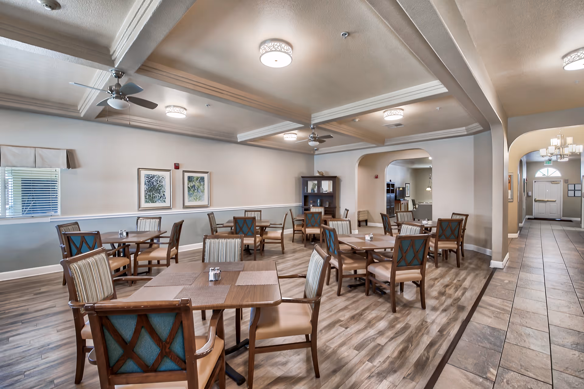 A spacious dining room in an assisted living facility with several wooden tables and chairs arranged neatly. The room features light-colored walls with two framed artworks, ceiling fans, and modern light fixtures. There is a tiled hallway leading to a door in the background.