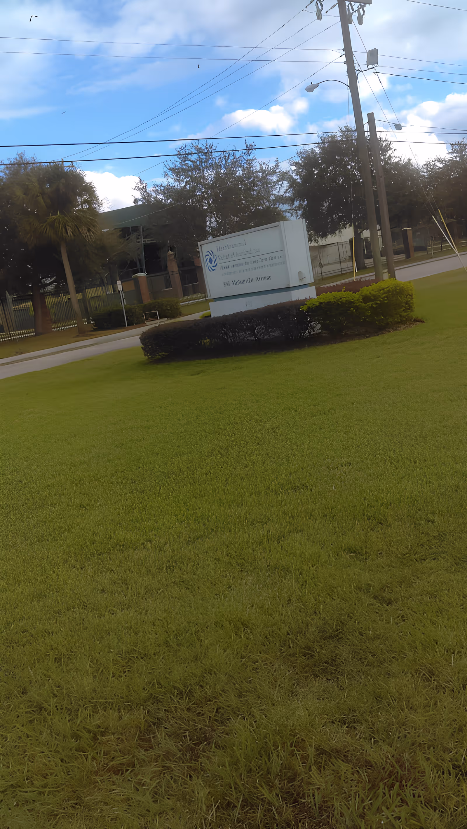 Outdoor view of a grassy area with a sign for Healthcare And Rehab Of Sanford, surrounded by bushes and trees, with a partly cloudy sky and power lines overhead.