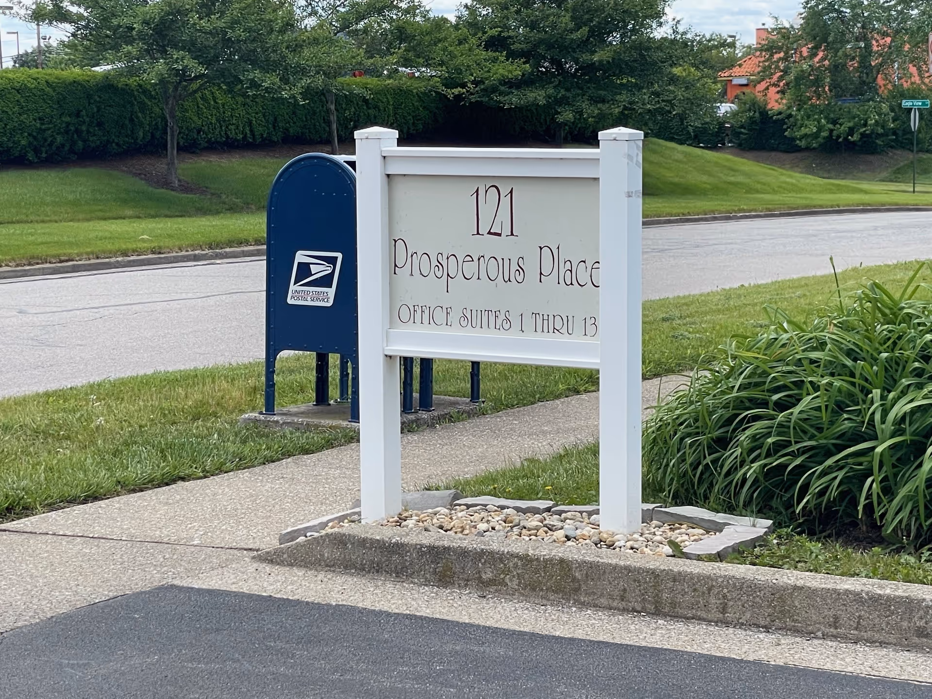 Street-side sign reads 121 Prosperous Place, Office Suites 1-13, with a mailbox and greenery.
