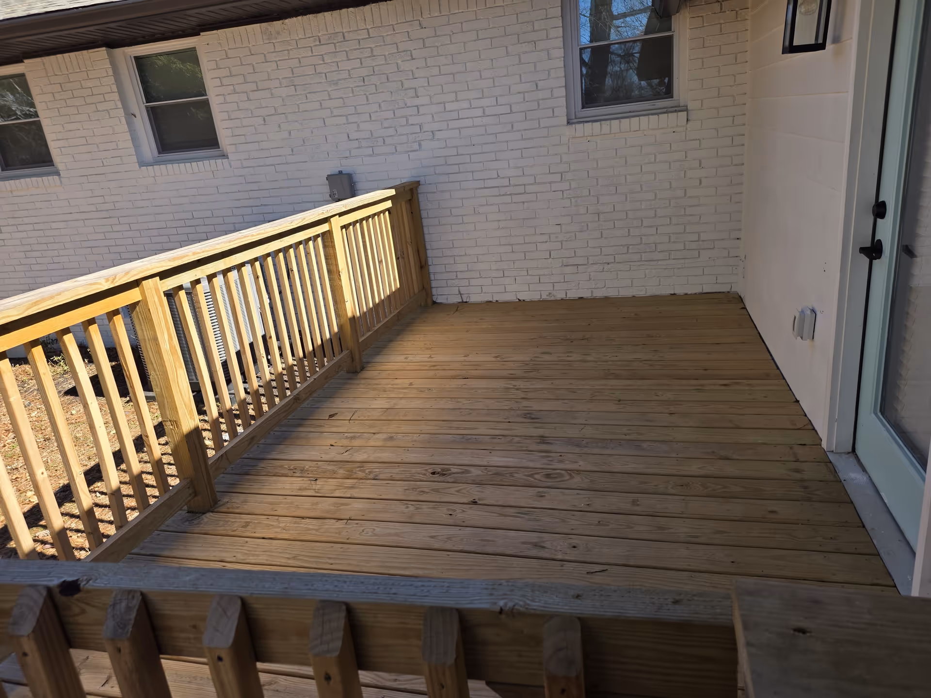 Exterior wooden deck with railing attached to a brick building beside a glass door.