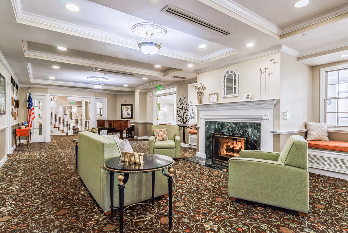 A spacious and well-lit living room area in Chapman Place featuring a green sofa and two matching armchairs arranged around a small round table with decorative letters spelling 'LOVE'. There is a fireplace with a green marble surround and a fire burning inside. The room has patterned carpet flooring, white walls, and ceiling lights. A piano is visible in the background near a staircase, and there are windows with cushioned seating along the wall.