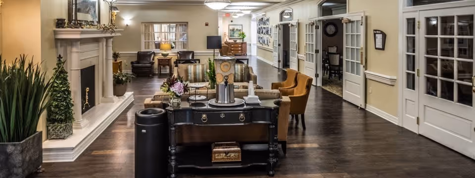 A spacious and well-lit senior living facility common area with wooden flooring, comfortable armchairs, a fireplace on the left, decorative plants, and a table with flowers and a decorative clock in the center. The hallway has multiple doors with glass panels and framed pictures on the walls.