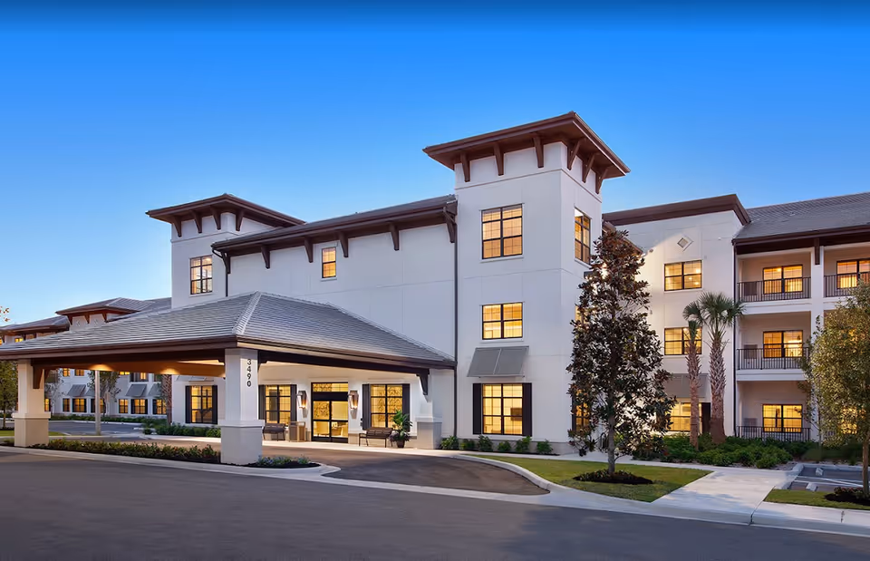 Exterior view of a modern senior living facility building named Seascape At Naples during dusk, featuring a covered entrance, multiple windows with warm interior lighting, landscaped greenery, and a clear blue sky.