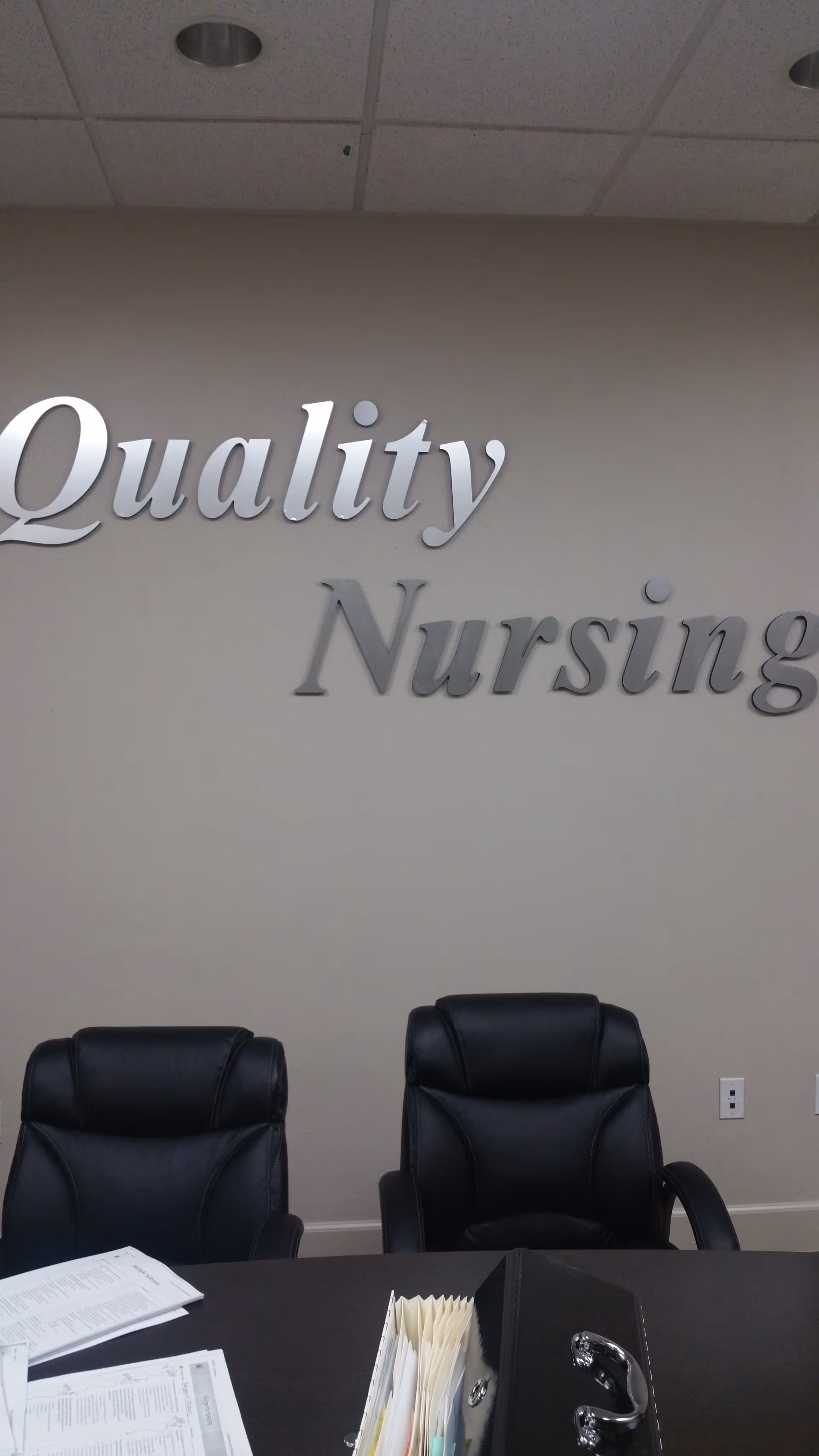 Interior wall features a large 'Quality Nursing' sign above a desk and black leather chairs.