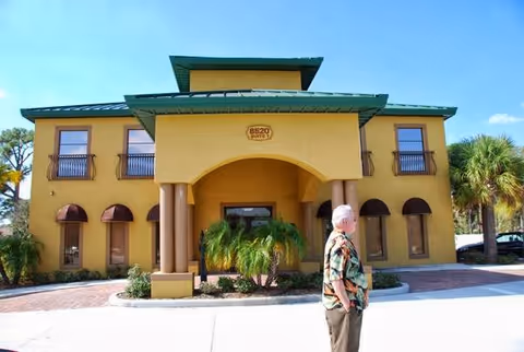 Yellow building with arched entrance and palm trees; a man stands in front.