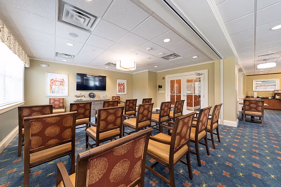 A well-lit meeting or presentation room with rows of wooden chairs featuring patterned upholstery facing a wall-mounted flat screen TV. The room has beige walls, a blue carpet with a floral pattern, framed artwork on the walls, and a sideboard with decorative items. There are double glass doors and a window with a valance allowing natural light in.