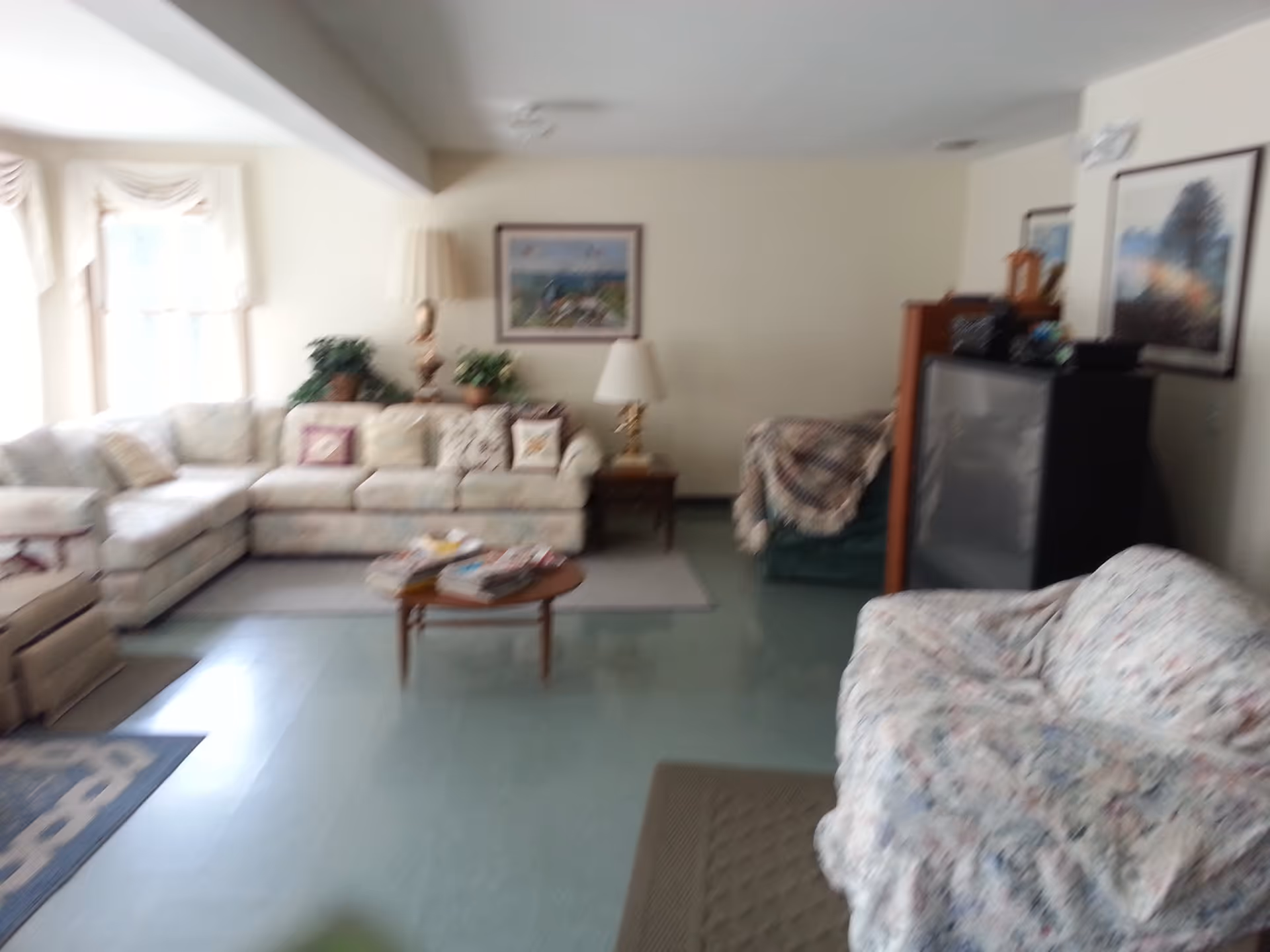 A living room with multiple upholstered sofas and armchairs arranged around a small wooden coffee table with magazines on it. The room has light-colored walls, framed paintings, table lamps, and potted plants near the windows.