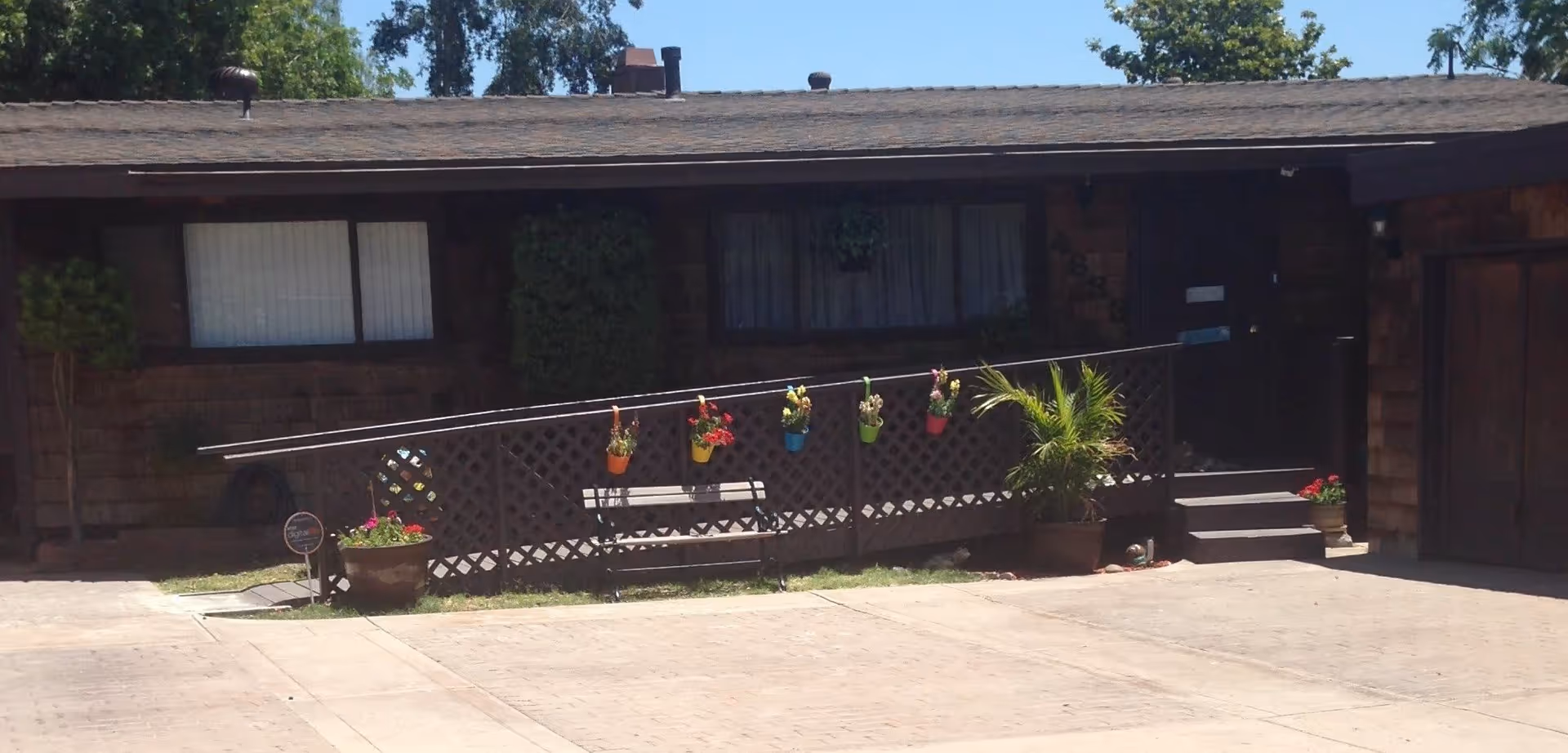 Front of a single-story building with a ramped porch, hanging and potted plants, and a bench.