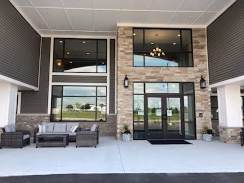 Covered entrance of a senior living building with glass double doors, large windows, stone facade, and outdoor seating.