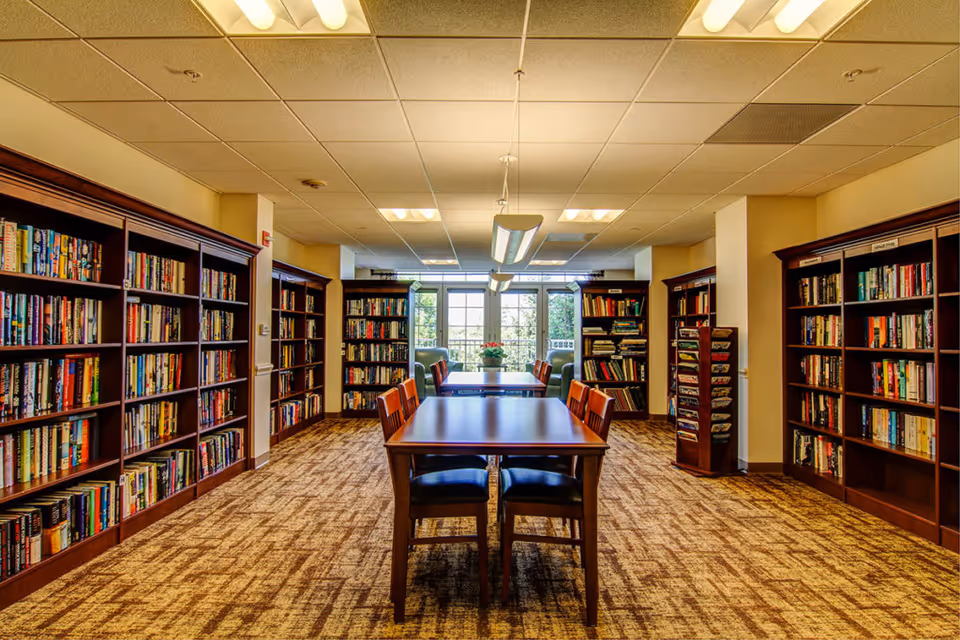 Well-lit library/reading room with a central table and chairs flanked by bookshelves and a window at the far end.
