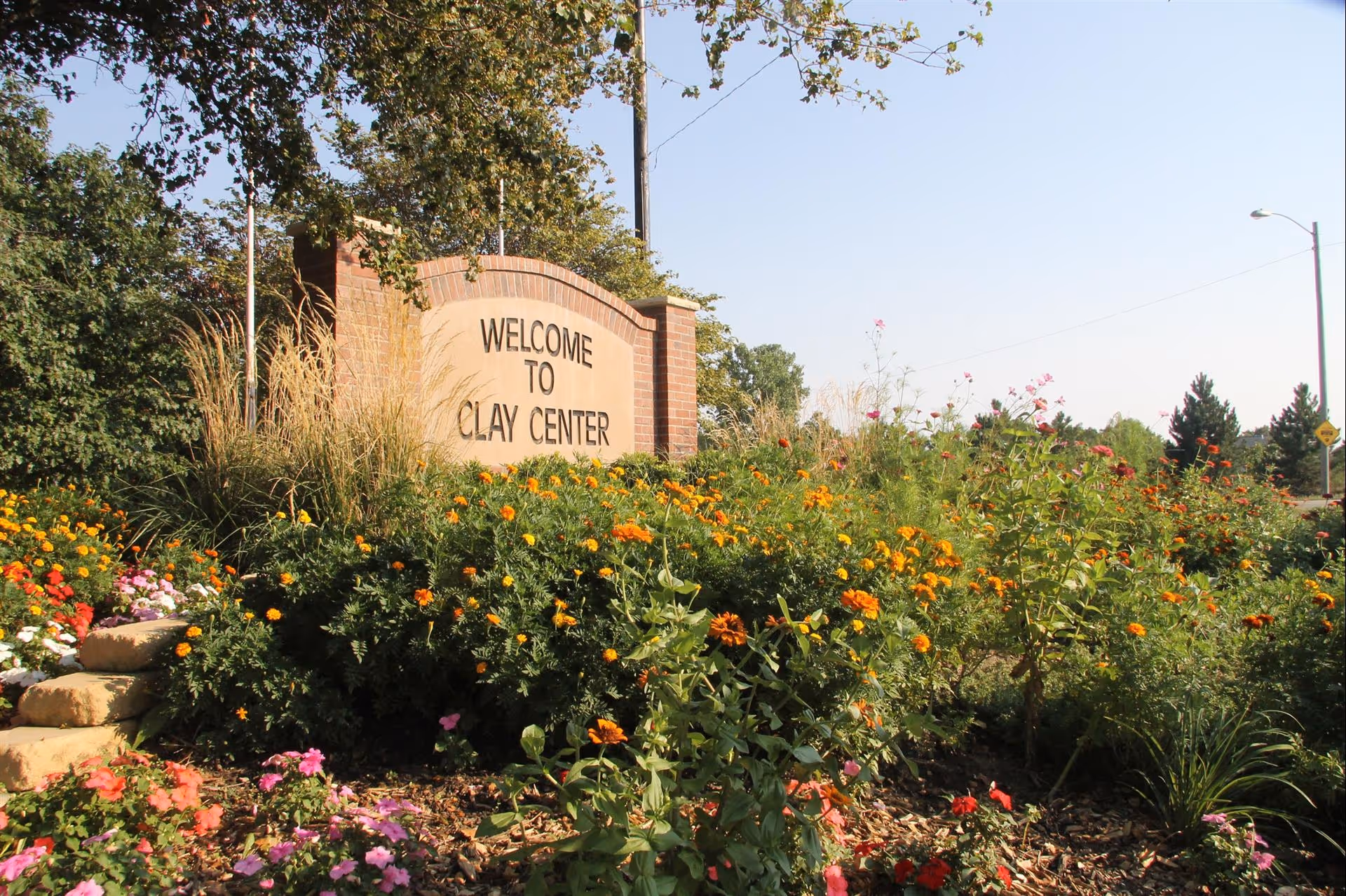A brick welcome sign reading "WELCOME TO CLAY CENTER" surrounded by colorful flowers and landscaping.