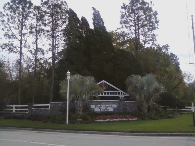 Landscaped entrance with a stone sign reading 'Fish Hawk Trails', flanked by palm trees and a white fence in front of tall trees.
