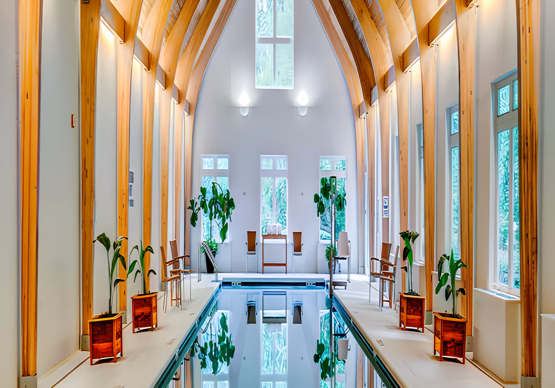 Indoor lap pool in a high-ceilinged room with arched wooden beams, tall windows, chairs, and potted plants.