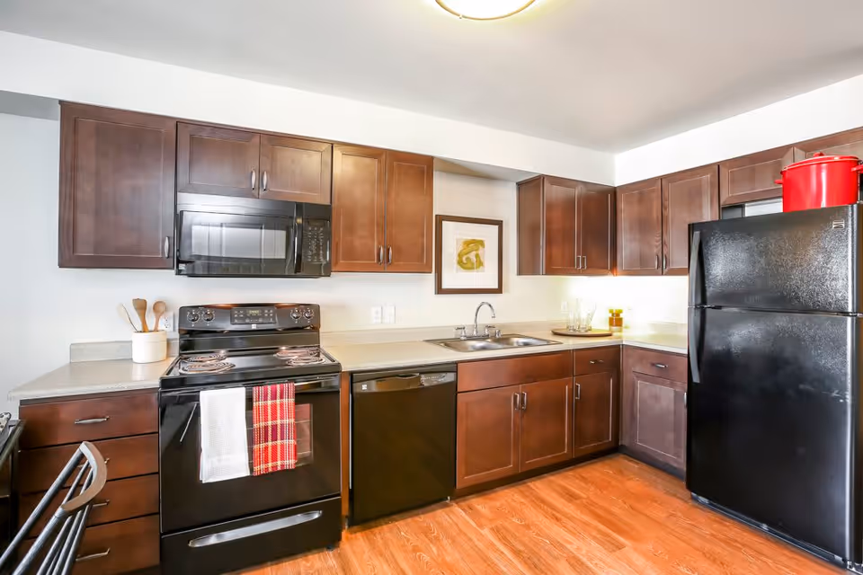 Modern kitchen with dark wood cabinets, black appliances including a stove, microwave, dishwasher, and refrigerator. The stove has two kitchen towels hanging on the handle. There is a double sink with a framed artwork above it, and a red pot on top of the refrigerator. The floor is wooden and the kitchen is well-lit.