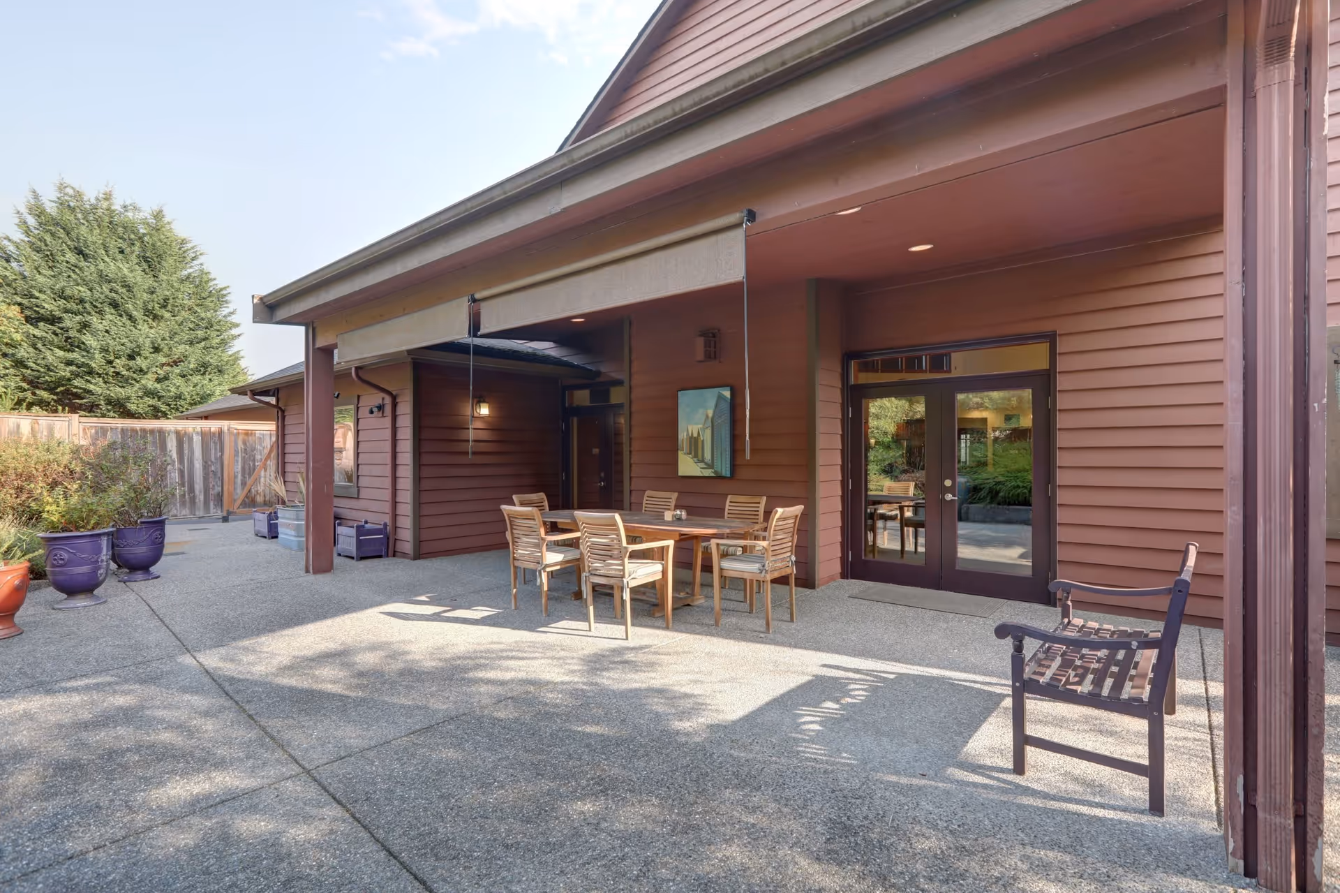 Outdoor patio area of a facility with a wooden table and six chairs under a covered section. There is a wooden bench to the right and several large potted plants along the left side. The building has brown siding and glass double doors leading inside.