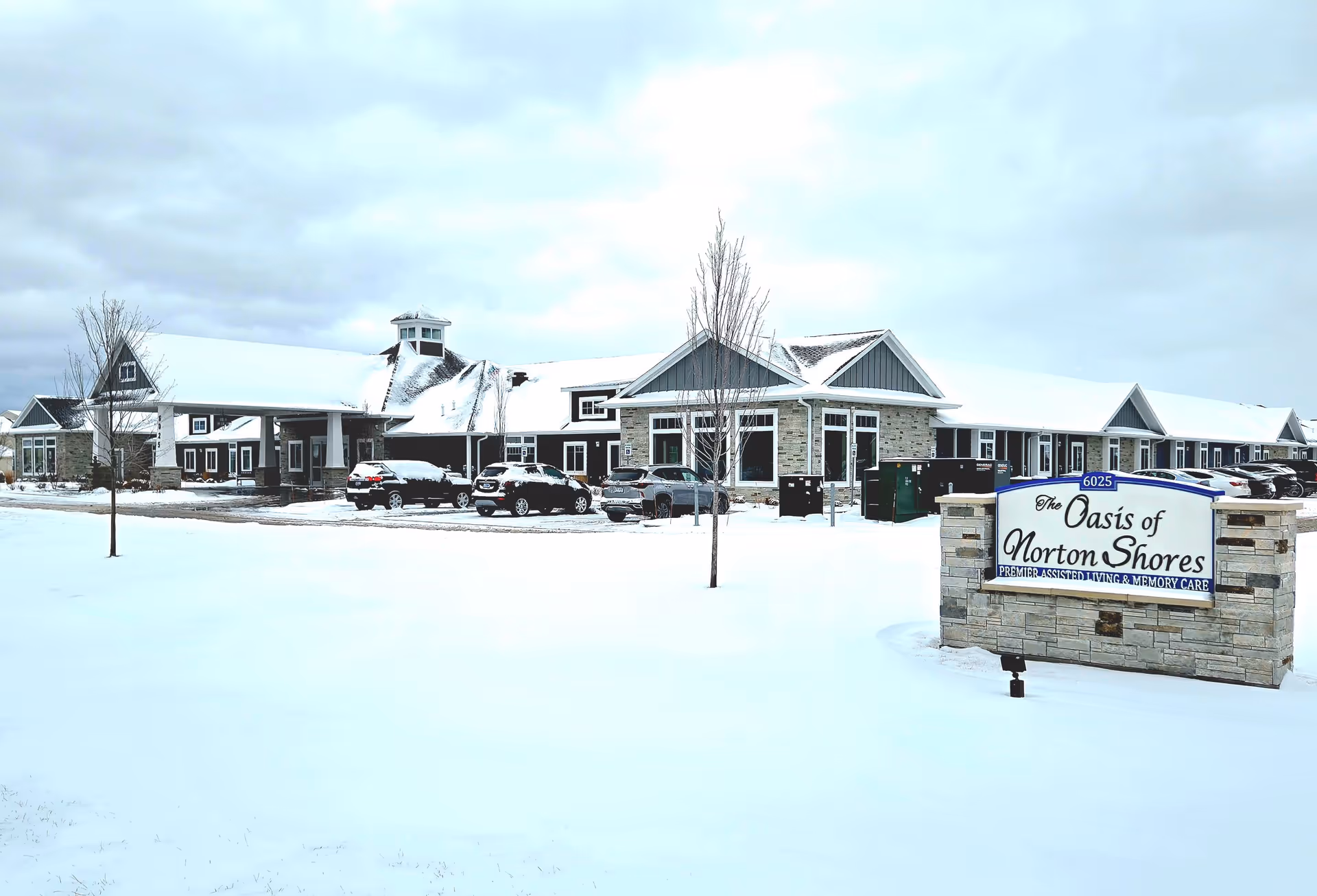 Exterior view of The Oasis of Norton Shores assisted living and memory care facility in winter, with snow covering the ground and roof, several parked cars, and a sign displaying the facility's name in front.