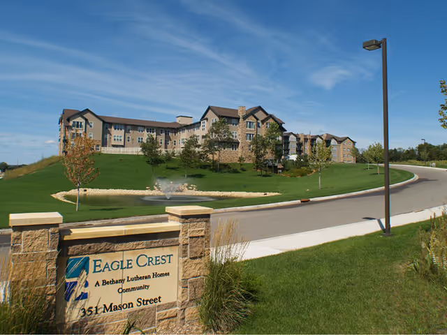 A multi-story Eagle Crest senior living building sits atop a grassy hill with a pond and fountain and a stone entrance sign in the foreground.