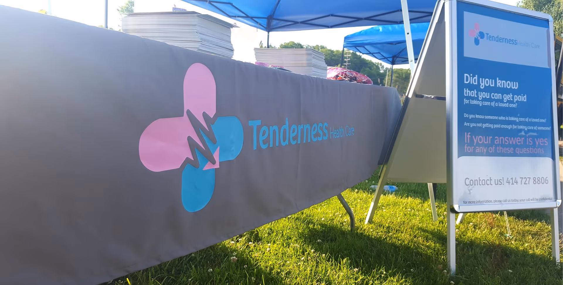 Outdoor event setup featuring Tenderness Health Care banner, blue canopies, and an informational sign.
