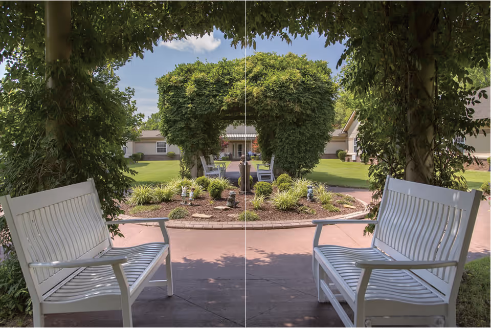 Two white benches facing each other under a pergola covered with green vines, overlooking a landscaped garden with trimmed bushes and a pathway leading to a building in the background under a clear sky.